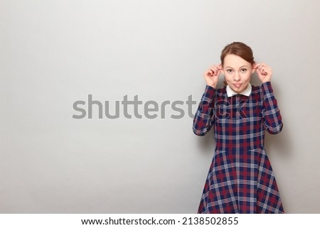 Studio portrait of funny happy young woman touching ears with hands, sticking out her tongue, fooling around and having fun, wearing checkered dress, standing over gray background, with copy space