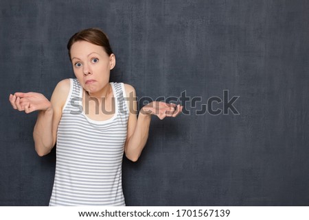 Studio portrait of funny perplexed girl wearing tank top, with stupid goofy face, grimacing, shrugging shoulders, raising hands, being bewildered, standing over gray background, copy space on right