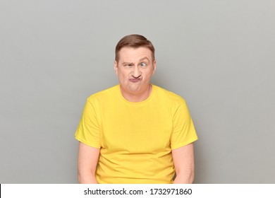 Studio portrait of funny puzzled blond mature man wearing yellow T-shirt, grimacing, making goofy crazy face, expressing perplexity and bewilderment, standing over gray background