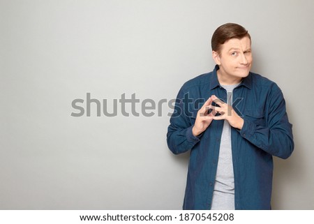 Studio portrait of funny thoughtful mature man wearing casual shirt, squinting, winking slyly, holding hands clasped, planning something profitable, standing over gray background, with copy space