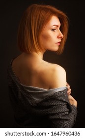 Studio Portrait Of A Ginger Girl With Freckles In A Dark Gray Shirt On A Black Background. With A Bare Shoulder, She Turned Her Back To The Frame And The Viewer