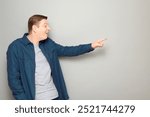 Studio portrait of goofy cheerful mature man pointing with finger at something funny, teasing somebody, laughing at joke, wearing blue shirt, standing sideways over gray background