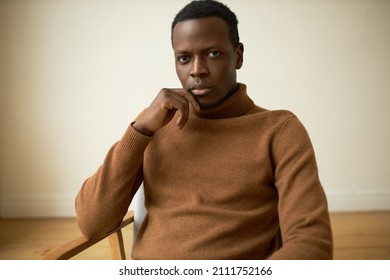 Studio portrait of handsome brutal serious and confident African American man in brown polo neck cashmere jumper, looking at camera with thoughtful face expression, resting chin on hand