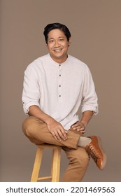 studio portrait of handsome Chinese man in his 30s smiling and sitting on a stool on neutral background Stock Photo