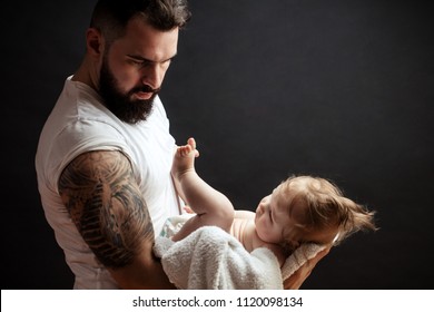 Studio Portrait Of Handsome Muscular Man In White T-short Holding On Tatooeed Hands Cute Little Baby Over Black Background With Copyspace. Family, Love And Happiness Concept.