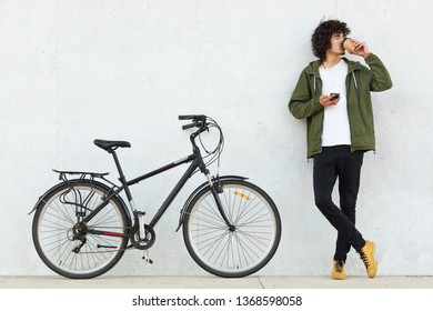 Studio portrait of handsome young Caucasian male with curly hair and little beard, drinking coffee and browsing internet using his smertphone, looking aside, standing at white wall near bicycle.