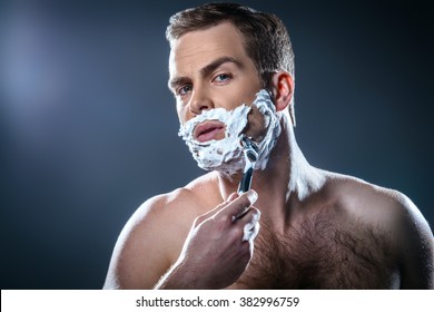 Studio portrait of handsome young man. Man with naked torso and shaving foam on face looking at camera and shaving