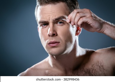 Studio Portrait Of Handsome Young Man. Clean Shaven Man With Naked Torso Looking At Camera And Tweezing Eyebrows