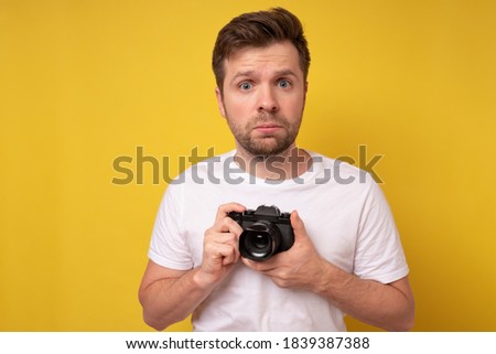 Studio portrait of handsome young man holding photocamera being confused in settings. Yellow blackground.