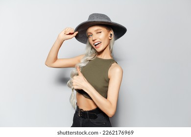 Studio portrait of happiness blonde girl with wireless earphones in ears, showing thumbs up, on white background. Wearing grey hat, green shirt and black jeans. - Shutterstock ID 2262176969