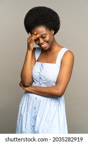 Studio portrait of happy African American young woman with afro hairstyle smiling looks embarrassed, smiling feels very confused. Shy millennial black girl dressed in blue dress isolated on gray wall