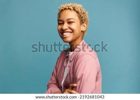 Studio portrait of happy african american girl with blond curls and nose ring standing in pink shirt with crossed hands laughing at joke on blue studio background. Positive emotions
