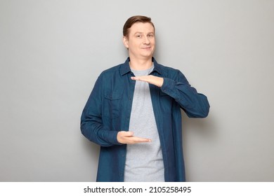 Studio Portrait Of Happy Blond Mature Man Showing Size Of Something, Holding Hands In Front Of Him With Place For Your Design, Smiling Cheerfully, Wearing Blue Shirt, Standing Over Gray Background