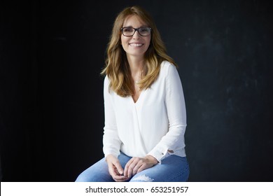 Studio Portrait Of A Happy Confident Middle Aged Woman Sitting At Black Background With Copy Space.