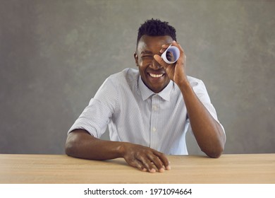 Studio Portrait Of Happy Curious Black Man Looking With One Eye Through Hole In Paper Roll. Cheerful Positive African American Guy In White Shirt Sitting At Desk Holding Document Scroll Like Telescope