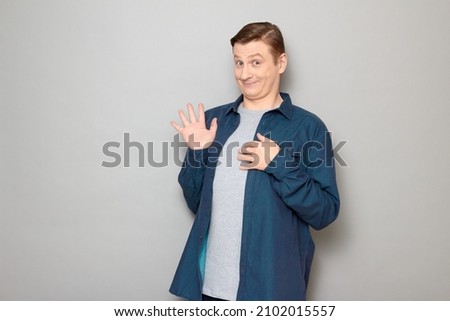 Studio portrait of happy funny blond mature man raising hand, waving palm, saying hello or goodbye, smiling joyfully, looking friendly and goofy, wearing blue shirt, standing over gray background