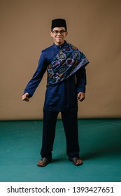 Studio Portrait Of A Happy, Handsome And Young Malay Asian Man In A Baju Melayu, Songkok Hat And Sash Dressed For Hari Raya Dancing In A Studio. He Is Smiling And Laughing As He Has Fun. 