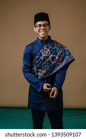 Studio Portrait Of A Happy, Handsome And Young Malay Asian Man In A Blue Baju Melayu, Songkok Hat And Patterned Sash Dressed For Hari Raya. He Is Smiling As He Poses For His Portrait Head Shot. 