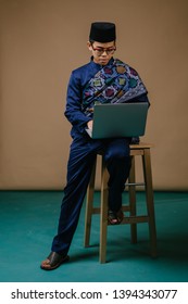 Studio Portrait Of A Happy, Handsome And Young Malay Asian Man In A Blue Baju Melayu, Songkok Hat With A Sash Sitting On A Chair And Working Or Studying On His Laptop Computer.