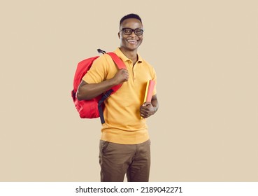 Studio portrait of happy school, college or university student. Handsome young African American man in polo shirt and glasses standing on beige background, holding books and backpack and smiling - Powered by Shutterstock