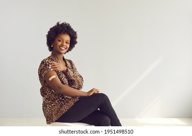 Studio portrait of happy young woman sitting on chair and showing her arm after getting vaccinated. Vaccination, safe Covid-19 vaccine, coronavirus immunity concept. Background with space for text