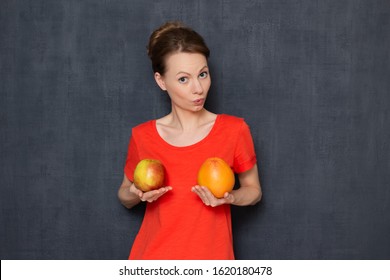 Studio portrait of impressed girl wearing T-shirt, holding big apple and grapefruit in hands over chest, demonstrating desirable bra size, having fun, fooling around, isolated over gray background