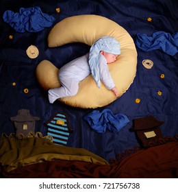 Studio Portrait Of Infant Baby Boy Wearing A Stocking Cap And Blue Pajamas, Sleeping On A Moon Shaped Pillow Against Textile Decoration Of A Night Village