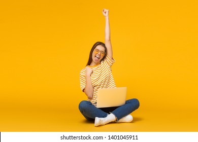 Studio portrait of joyful girl with laptop computer, sitting on a floor and celebrating, isolated on yellow background - Shutterstock ID 1401045911