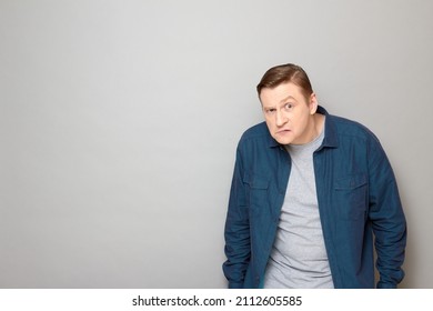 Studio portrait of mature man looking distrustfully and suspiciously with one eyebrow raised, with frowning face, wearing casual blue shirt, standing hunched over gray background, with copy space