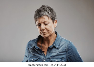 Studio portrait of a mature woman with short gray hair and a denim shirt, looking down with closed eyes, conveying feelings of sadness, disappointment, and negativity - Powered by Shutterstock