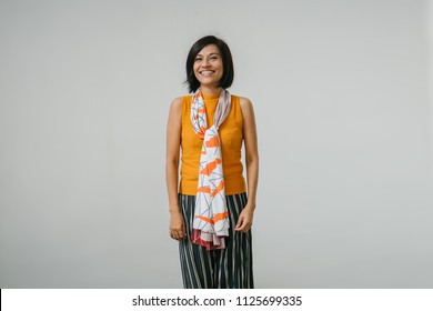 Studio Portrait Of An Older And Attractive Chinese Asian Woman Against A White Backdrop. She Is Smiling And Is Wearing An Orange Sweater, A Dark Dress And Has A Scarf Draped Over Her Neck. 