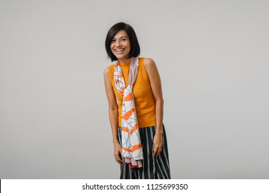 Studio Portrait Of An Older And Attractive Chinese Asian Woman Against A White Backdrop. She Is Smiling And Is Wearing An Orange Sweater, A Dark Dress And Has A Scarf Draped Over Her Neck. 