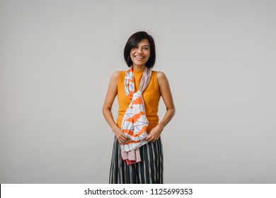 Studio Portrait Of An Older And Attractive Chinese Asian Woman Against A White Backdrop. She Is Smiling And Is Wearing An Orange Sweater, A Dark Dress And Has A Scarf Draped Over Her Neck. 