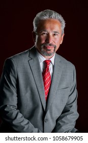 Studio portrait of an older, handsome, latino business man.  He is leaning in front of a dark red background. The light is dramatic.