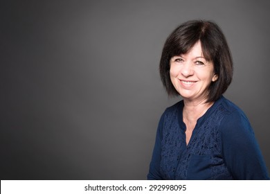 Studio Portrait Of An Older Woman With Dark Hair Wearing A Blue Shirt Against A Gray Background.  Horizontal Format.