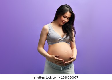 Studio Portrait Of Pregnant Caucasian Woman With Long Dark Hair. Happy Future Mom Posing With Hands On Naked Belly Over Purple Background