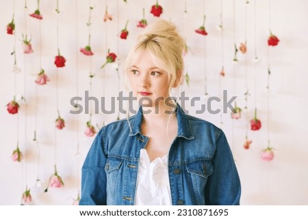 Studio portrait of pretty young teenage 15 - 16 year old girl wearing denim jacket, posing on white background with hanging flowers, beauty and fashion concept