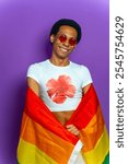 Studio portrait with purple background of a transgender latin man wrapped with lgbt flag smiling at camera