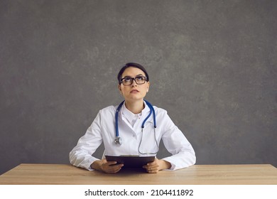 Studio Portrait Of A Serious Female Doctor Thinking About Something. Young Woman In Glasses And White Medical Lab Coat Looking Up Sitting At An Office Desk At Hospital