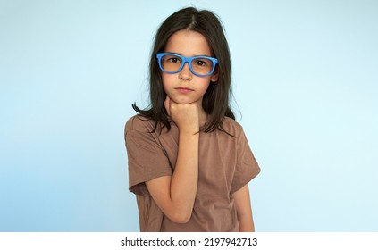 Studio Portrait Of A Serious Kid Wearing Blue Eyeglasees Looking At The Camera Posing Over Light Blue Studio Background. A Thoughtful Little Girl Thinking About Something.