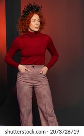 Studio Portrait Of A Serious Woman 35-40 Years Old With Red Hair And A Crown On Her Head In The Form Of Snakes On A Colored Background, Hands In Pockets.