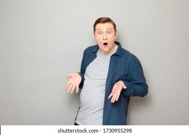 Studio Portrait Of Shocked Blond Mature Man With Widened Eyes And Open Mouth, Wearing Blue Shirt, Having Overweight, Being Surprised With His Big Stomach And Fat Body, Standing Over Gray Background