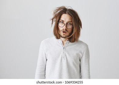 Studio portrait of shocked messy guy in glasses being surprised and displeased, arguing with colleague, feeling tired after doing hard project alone, standing against gray background bothered