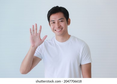 Studio Portrait Shot Of Asian Happy Young Handsome Smart Teenager Teen Guy Model In Casual Clothing Smiling Show Teeth Look At Camera Waving Hand Greeting Say Hello Hi Of Goodbye On White Background.