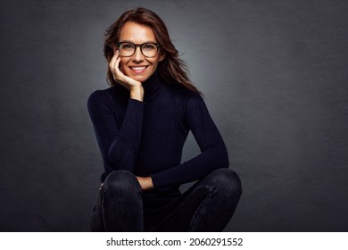 Studioaufnahme einer attraktiven Frau mittleren Alters, die einen Schildkrötenpullover trägt und lacht, während sie auf isoliertem dunklem Hintergrund sitzt. Kopiert Platz.