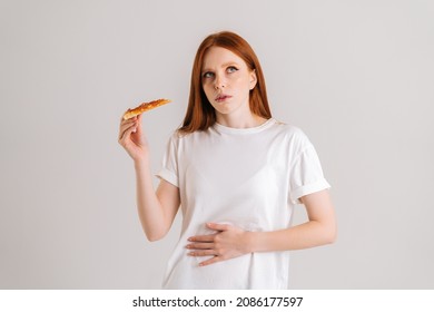 Studio Portrait Of Sick Unhappy Young Woman Feeling Pain In Stomach After Eating Pizza, Suffering Abdomen Ache Looking Away, Standing On White Isolated Background, Holding Pizza Slice.