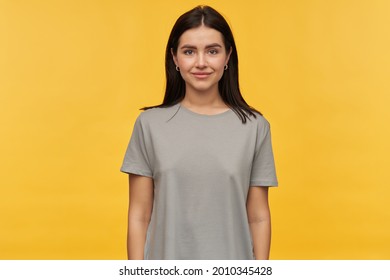 Studio Portrait Of Smiling Beautiful Young Woman With Dark Hair In Gray Tshirt Standing And Lookign At Camera Over Yellow Background