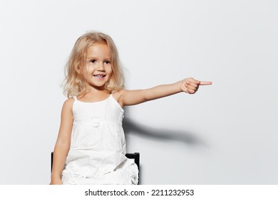 Studio Portrait Of Smiling Child Girl With Blonde Hair Pointing Index Finger Away On White Background.