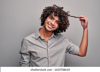 Studio Portrait Of Smiling Eastern Hadsome Boy Holding A Strand Of His Hair In Hand Posing On Gray Isolated Background. Arabian Man Shows His Healthy Beautiful Curly Hair