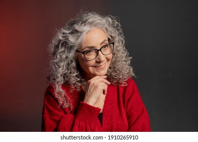 Studio Portrait Of A Smiling Elderly Woman 60-65 Years Old In A Red Sweater And Glasses, Gray Curly Long Hair, On A Colored Background. Concept: Stylish Pensioners Of Model Appearance, Active Life.